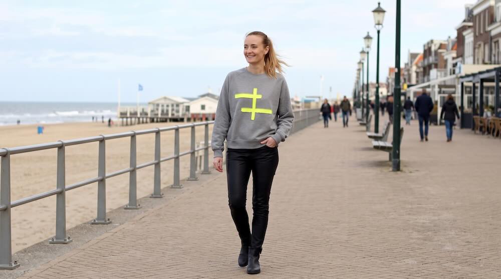 Vrouw in de grijs melange sweater Fluor Yellow Cross met neongeel kruis, gedragen op een zwarte leren broek en zwarte enkellaarzen op een strandboulevard