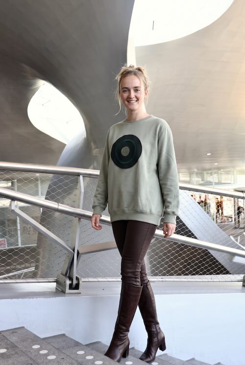 Vrouw in de lange saliegroene sweater Sage Circles met donkergroene cirkels, gedragen op een strakke donkerbruine broek en hoge bruine laarzen in de stationshal van Arnhem Centraal