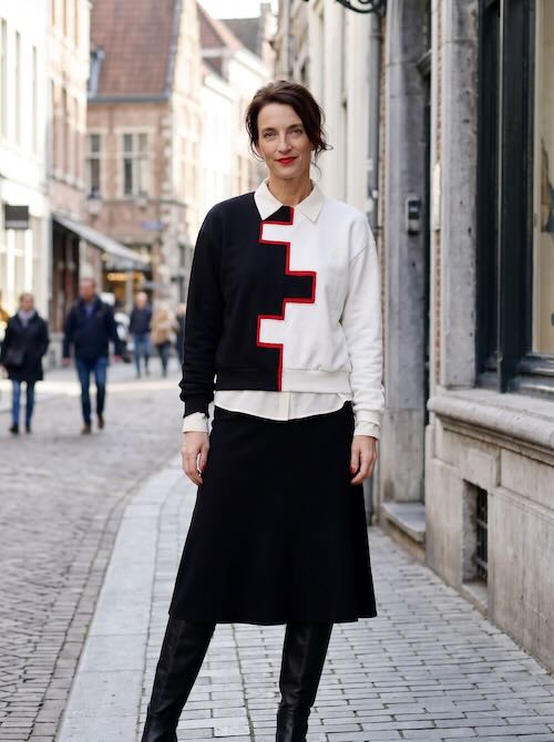Vrouw in een zwart-witte sweater met rode getrapte lijn, gedragen over een witte blouse op een zwarte midirok en zwarte laarzen