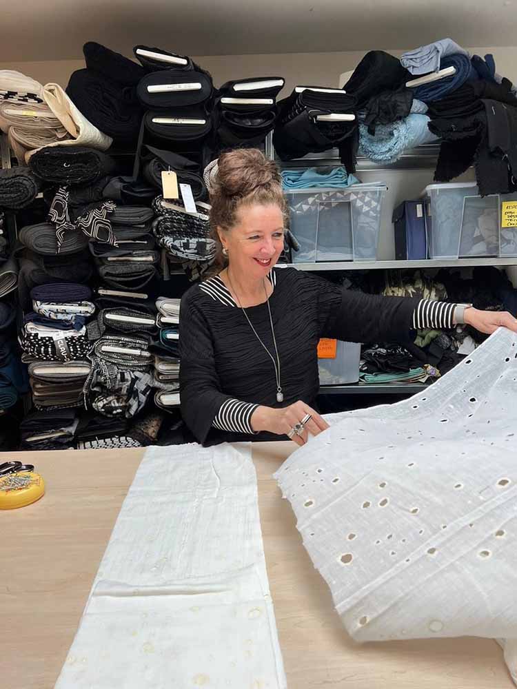 Een professionele foto van ontwerper Claudia Träumer in haar gedetailleerde Arnhem Modekwartier atelier voor CLAU-D. Ze staat achter een grote houten snijtafel en presenteert een prachtige, teal zijde-wol mix stof terwijl ze omringd is door planken en displays met een zorgvuldig samengestelde collectie van duurzame stoffen (zoals wol, linnen en gecertificeerd biologisch katoen). Op de achtergrond is een groot, karakteristiek boograam te zien met uitzicht op het historische Modekwartier in Arnhem.